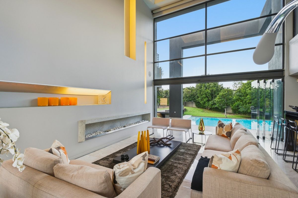 Spacious living room featuring double-volume ceilings, floor-to-ceiling glass sliding doors, integrated fireplace, and seamless flow to the outdoor pool area.