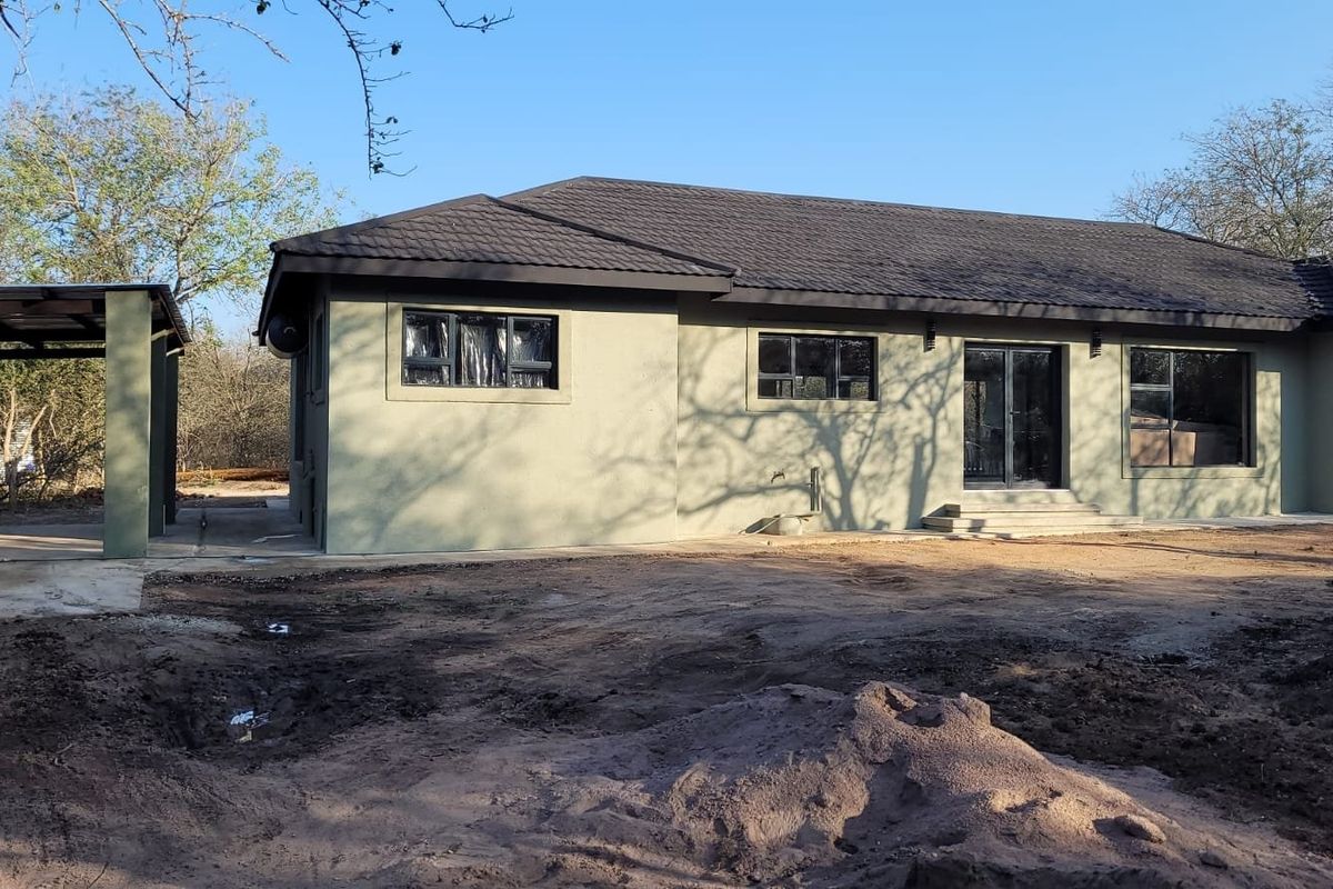 Rear view of a single-story house featuring a dark tiled roof, neutral exterior walls, and a covered carport structure with an unfinished garden area.