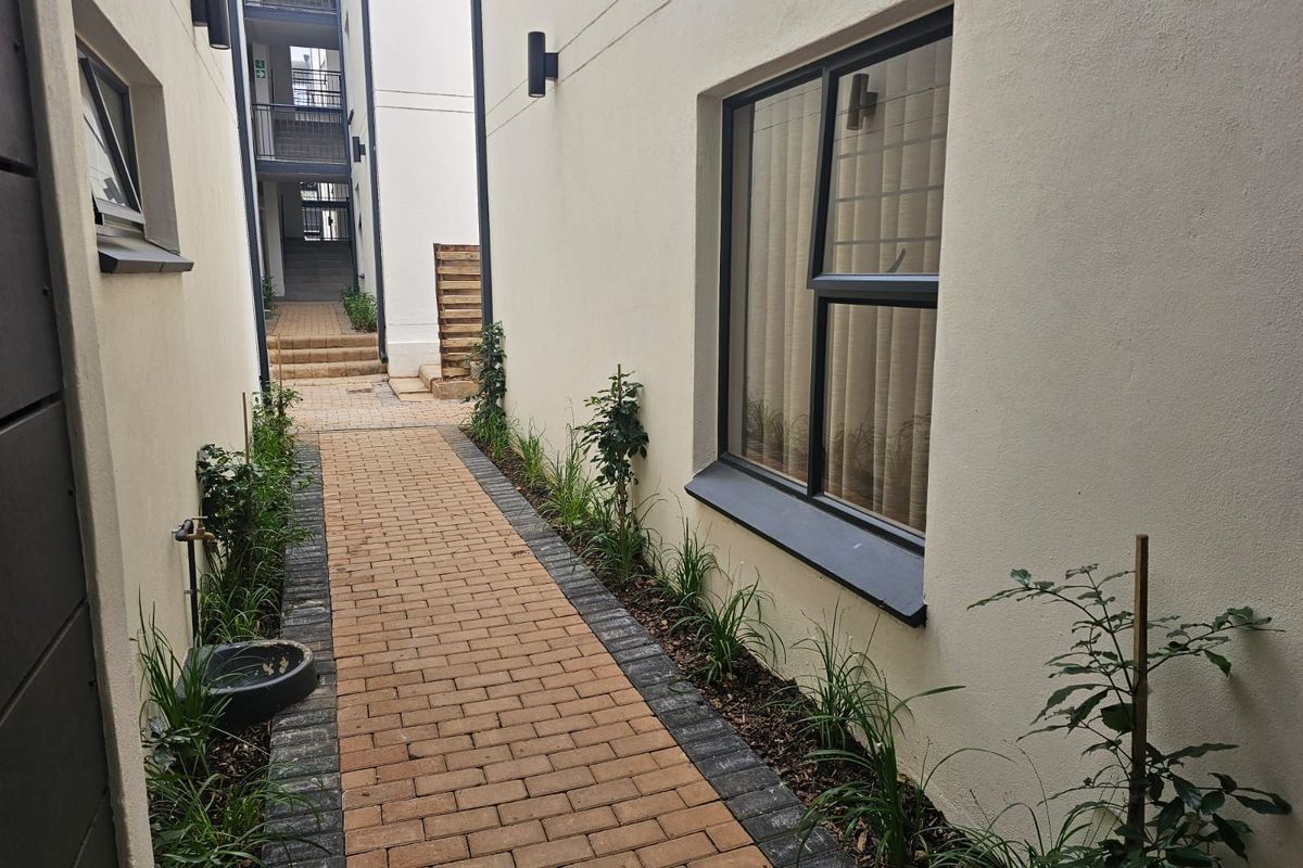 A paved side walkway between apartment building sections, featuring landscaped garden beds, exterior wall lighting, and a functional outdoor water tap.