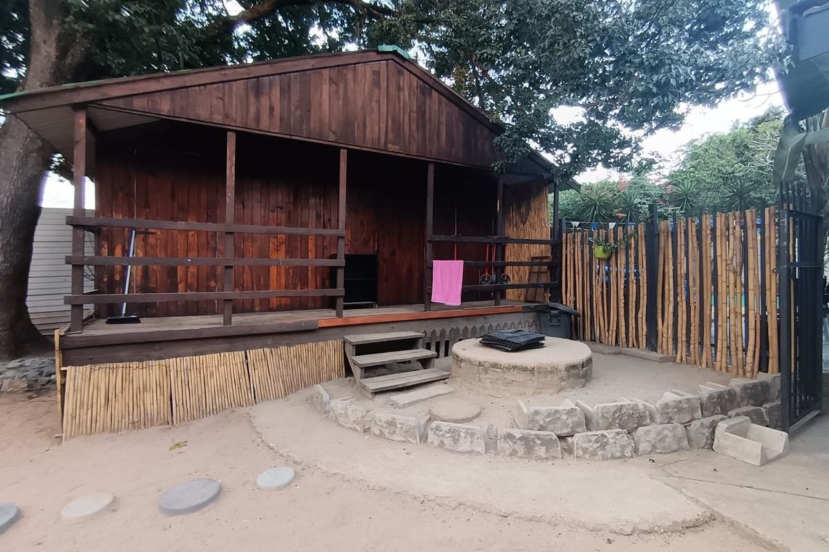 A rustic wooden cabin featuring a covered front porch, elevated timber decking, and an adjacent circular stone fire pit area in a garden setting.