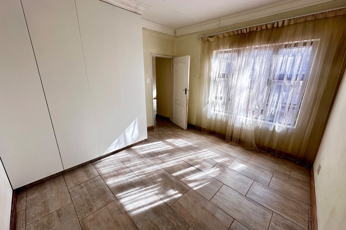 A bright, empty bedroom featuring tiled flooring, built-in cupboards, and large windows with natural light.