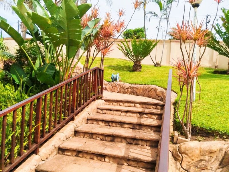 A landscaped garden featuring stone-clad stairs with wooden railings, leading up to a manicured lawn area enclosed by a perimeter wall.