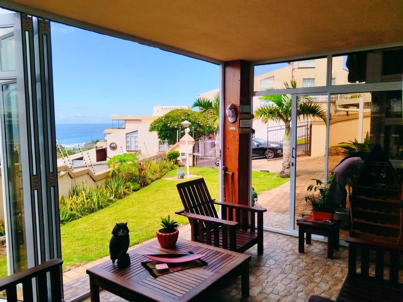 A covered patio area featuring tiled flooring and large glass sliding doors, offering views of the garden and the ocean beyond.
