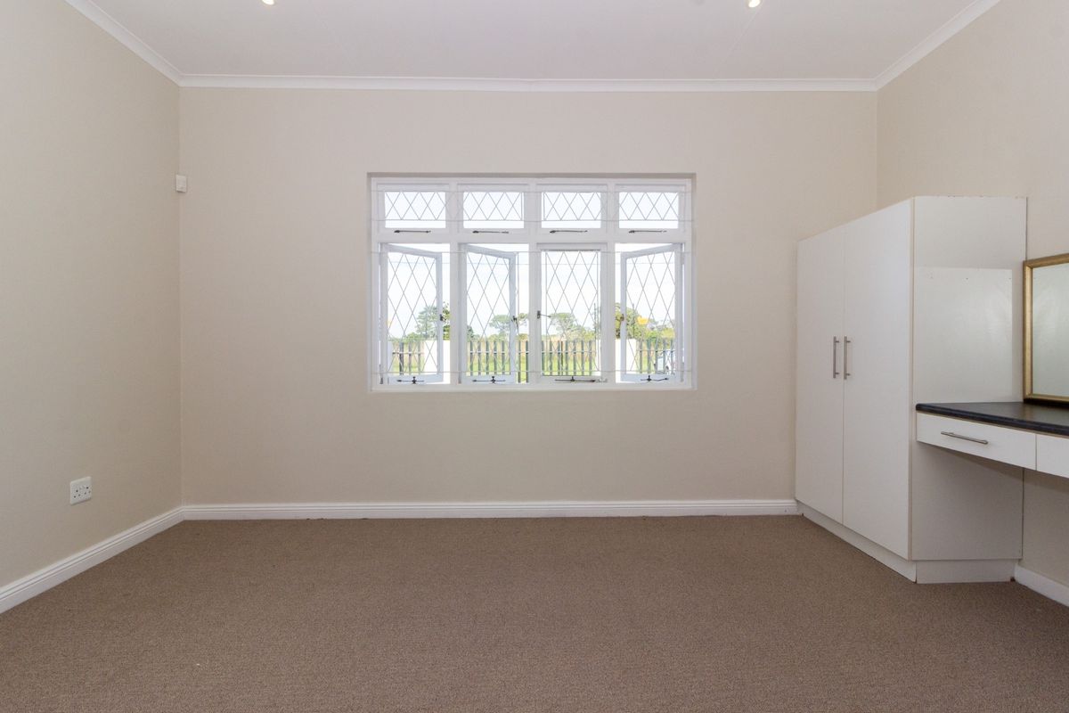 A clean, neutral bedroom featuring fitted built-in cupboards, a vanity unit, and large windows with security bars, finished with carpeted flooring.
