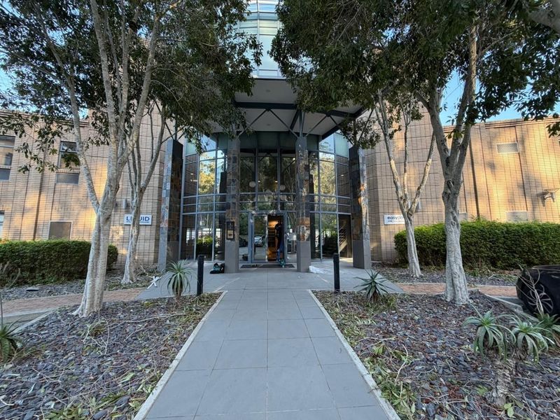 Front entrance of a modern office building featuring a glass facade, stone-clad columns, and a paved pedestrian walkway.