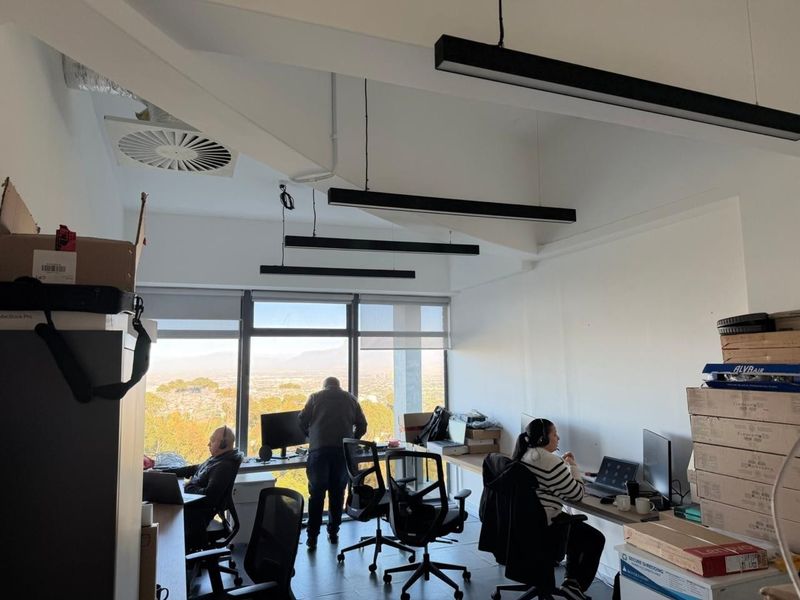 A bright open-plan office space featuring modern suspended linear lighting, large windows with panoramic views, and integrated ceiling air conditioning.