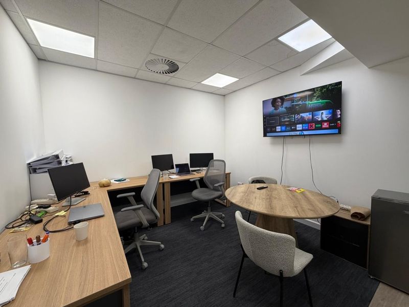 A functional office space featuring a suspended ceiling with recessed lighting, air conditioning, and carpeted flooring, equipped with a wall-mounted television.