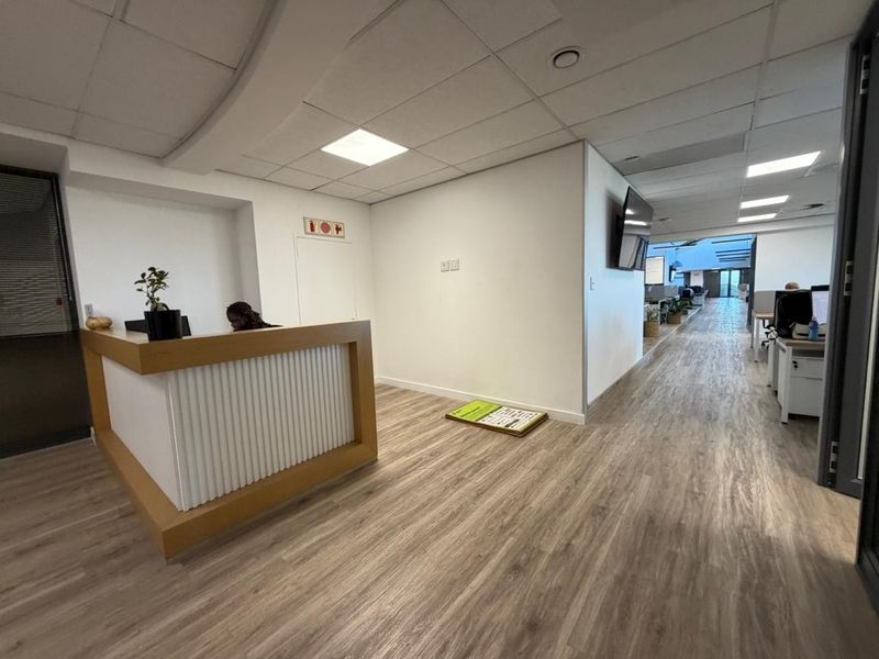 Modern office reception area featuring a built-in desk, suspended ceiling with recessed lighting, and wood-look laminate flooring leading into an open-plan workspace.