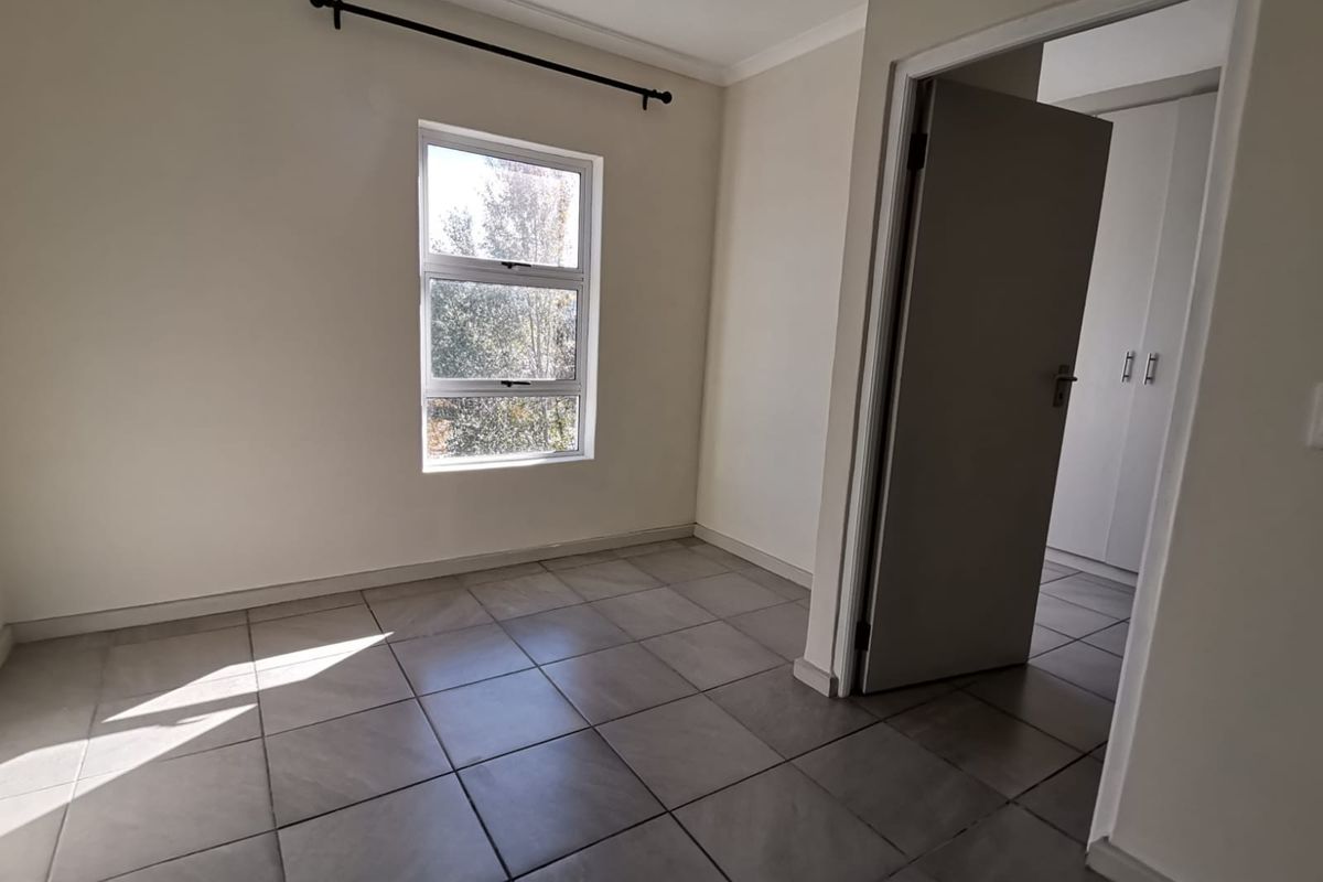 A bright, empty bedroom featuring neutral tiled flooring, a window with natural light, and access to a hallway with built-in cupboards.