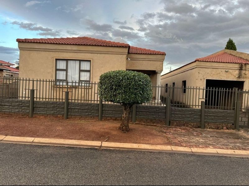 Front exterior view of a single-story house featuring a tiled roof, stucco facade, and secure perimeter fencing with a gated entrance.