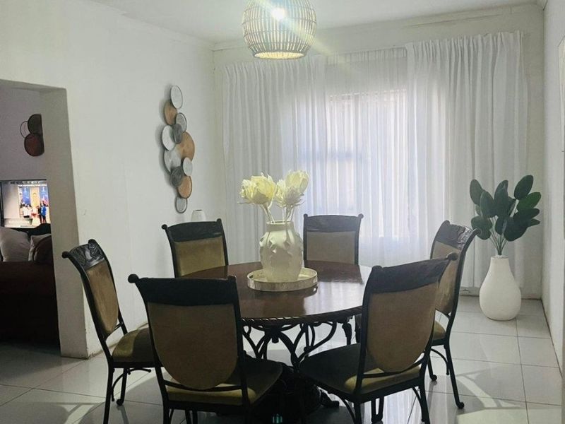 A bright dining area featuring tiled flooring, a central pendant light fixture, and large windows dressed with floor-to-ceiling curtains.