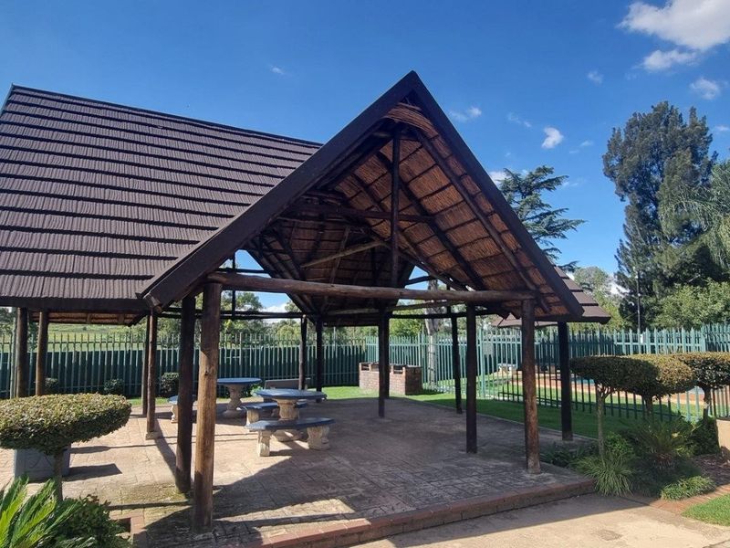 A covered outdoor entertainment area featuring a thatched roof structure, built-in brick braai, and paved flooring, situated within a landscaped garden setting.