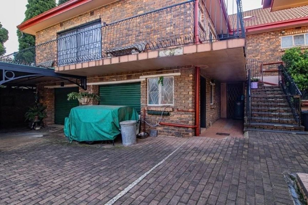 Exterior view of a multi-level brick residence featuring a paved driveway, covered parking area, external staircase, and balcony with metal security railings.