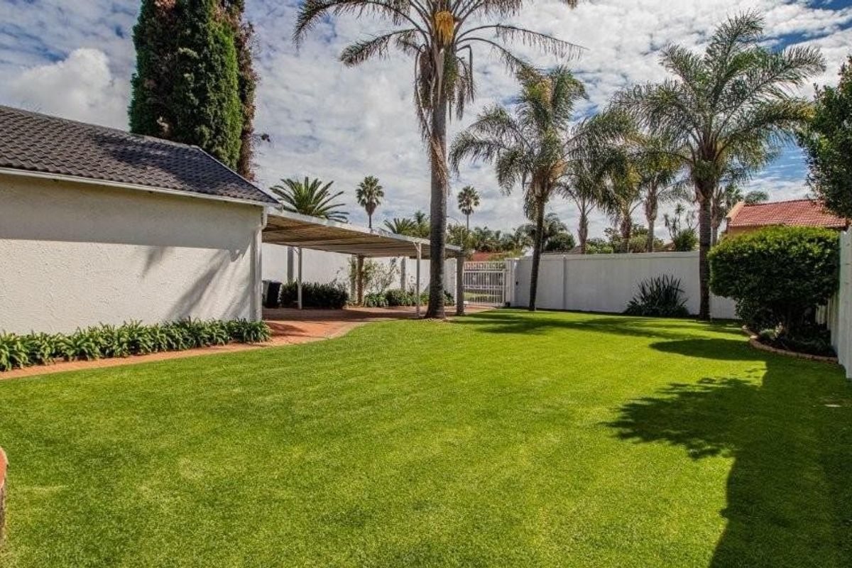 A well-maintained rear garden featuring a lush green lawn, paved pathways, a covered carport area, and perimeter boundary walls.