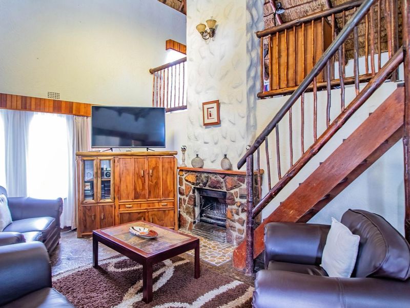 Spacious living room featuring a double-volume thatched ceiling, stone-clad fireplace, and a rustic timber staircase leading to an upper mezzanine level.