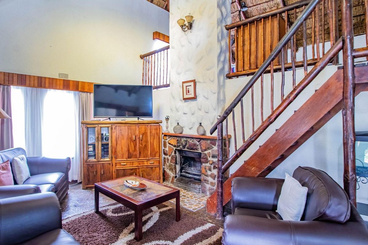 Spacious living room featuring a double-volume thatched ceiling, stone-clad fireplace, and a rustic timber staircase leading to an upper mezzanine level.
