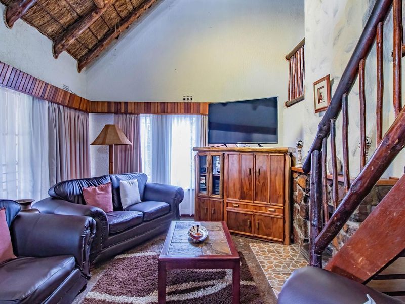 A rustic living room featuring a high vaulted thatched ceiling, exposed timber beams, and a stone-clad fireplace with a wooden staircase.
