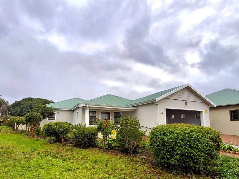 Front exterior view of a single-story residential house featuring a green metal roof, paved driveway, and attached garage, set within a landscaped garden.