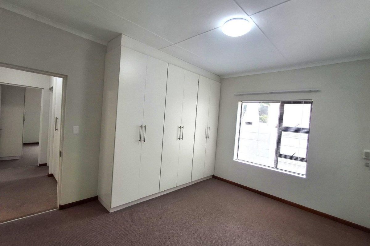 A clean, neutral bedroom featuring built-in white cabinetry, carpeted flooring, and a window with fitted blinds.