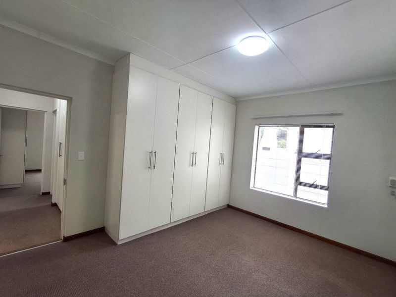 A clean, neutral bedroom featuring built-in white cabinetry, carpeted flooring, and a window with fitted blinds.