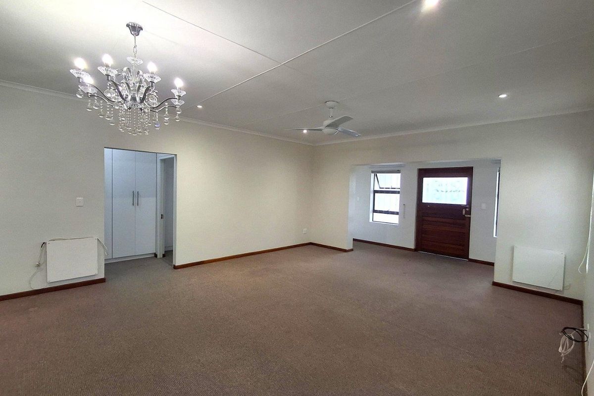 Spacious living area featuring neutral carpeting, a decorative chandelier, ceiling fan, and built-in wall heaters, with an adjacent hallway and exterior access door.