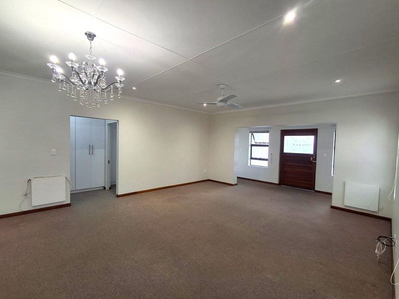 Spacious living area featuring neutral carpeting, a decorative chandelier, ceiling fan, and built-in wall heaters, with an adjacent hallway and exterior access door.