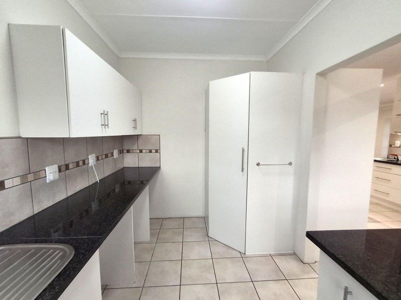 A functional scullery area featuring dark granite countertops, white cabinetry, tiled splashbacks, and tiled flooring, with a view into the adjacent kitchen.