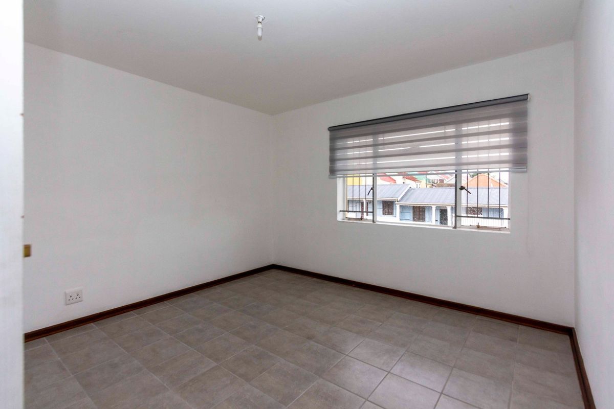 A clean, empty bedroom featuring neutral floor tiles, white walls, and a window fitted with a modern roller blind and security bars.