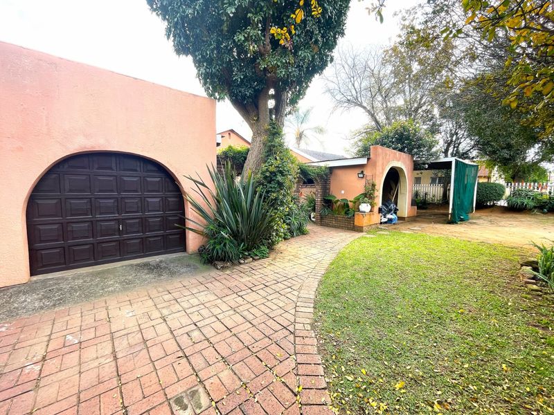Front exterior view featuring a paved driveway, arched garage door, and landscaped garden area with mature trees.