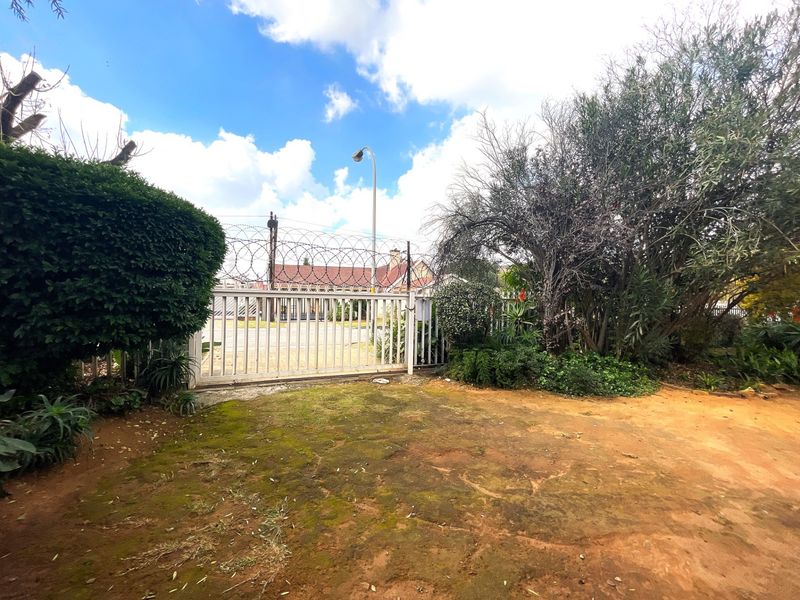 A view of the property entrance featuring a metal security gate topped with razor wire, set within a garden area with unpaved ground.