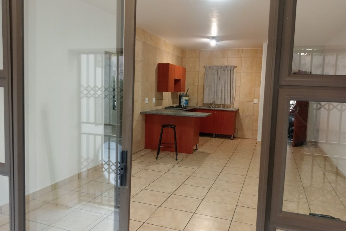 An open-plan kitchen featuring tiled flooring, built-in cabinetry with laminate countertops, a built-in hob, and a stainless steel sink, viewed through a sliding door.