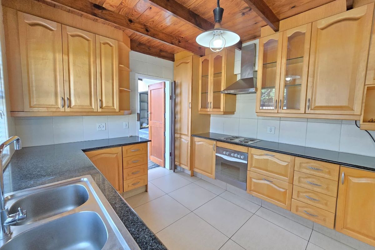 A functional kitchen featuring light wood cabinetry, dark granite countertops, a stainless steel extractor fan, and a double basin sink under a timber-beamed ceiling.