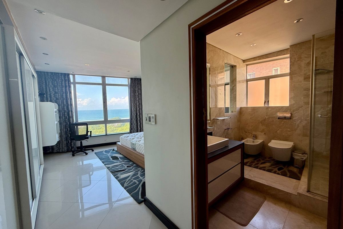 A modern bedroom suite featuring floor-to-ceiling windows with ocean views, tiled flooring, and an en-suite bathroom with marble-style wall tiling and contemporary fixtures.