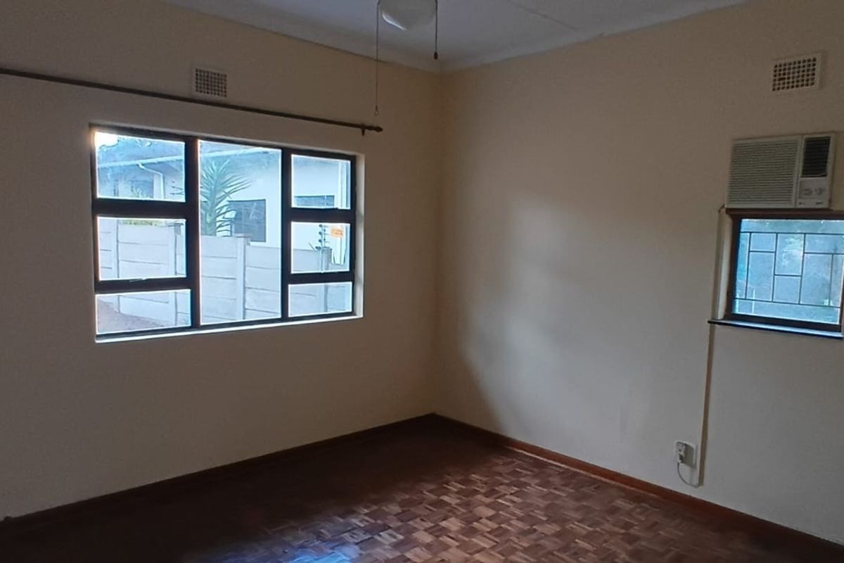 A vacant bedroom featuring parquet flooring, large windows with security bars, and a wall-mounted air conditioning unit.
