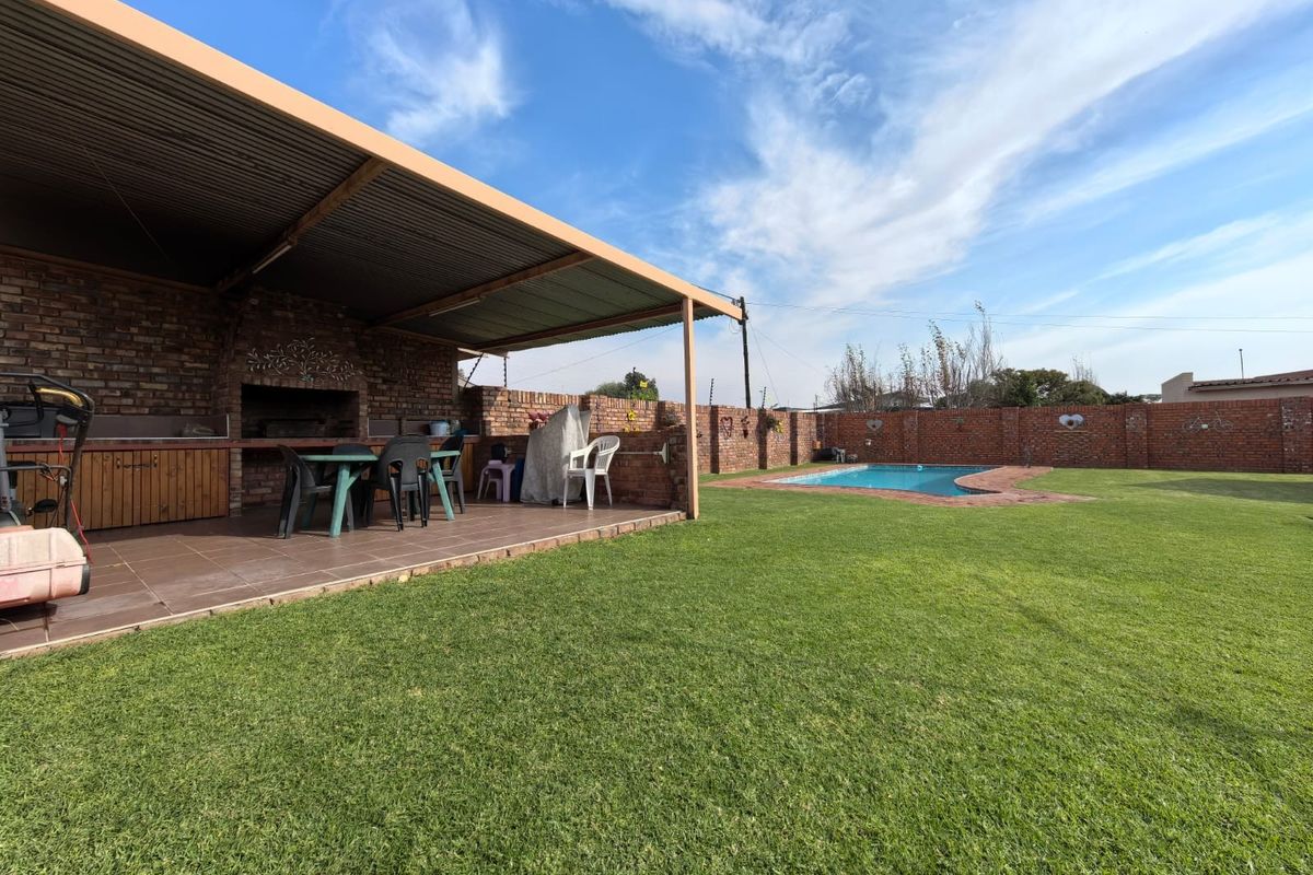 Spacious backyard featuring a covered patio with built-in braai area, a swimming pool, and a well-maintained lawn enclosed by brick perimeter walls.