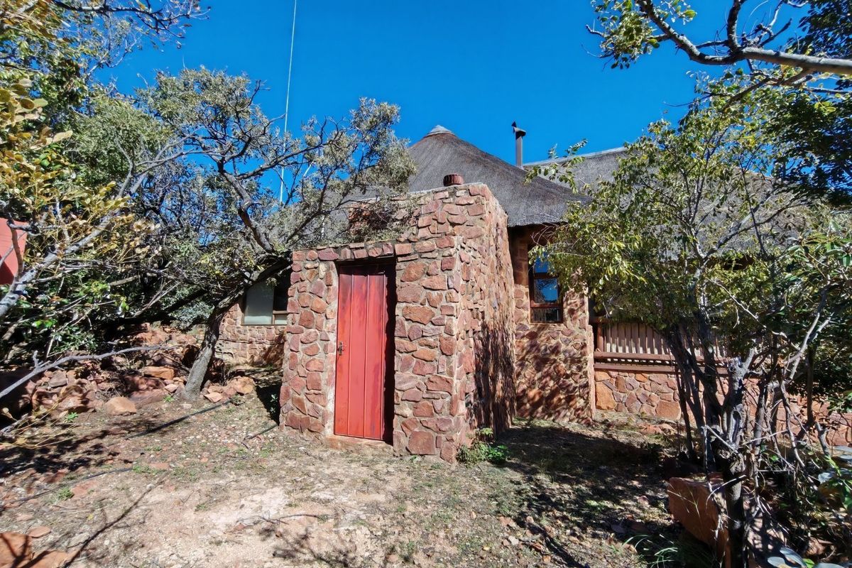 A rustic exterior view of a stone-clad house featuring a traditional thatched roof and a wooden entrance door set within a natural garden.