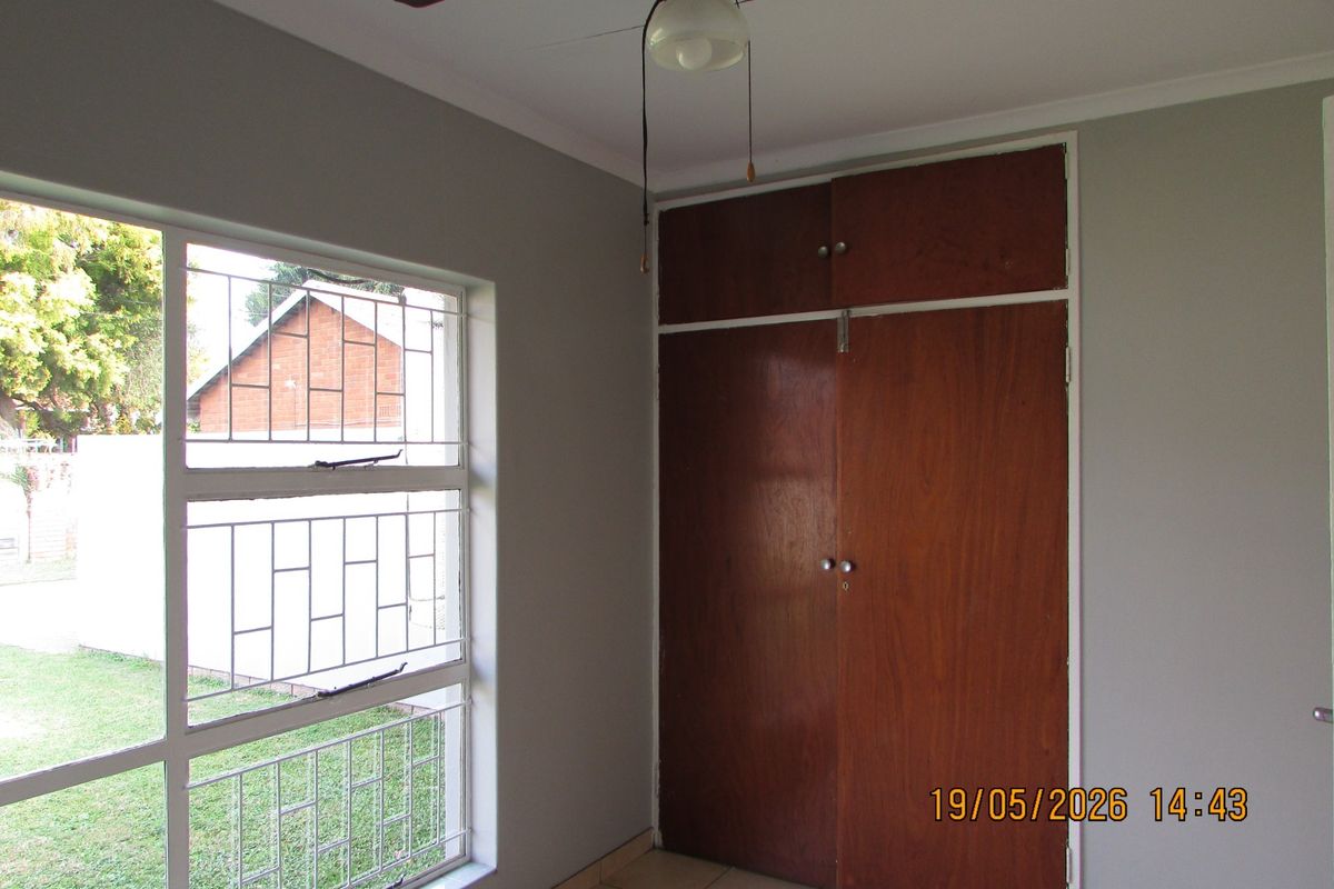 A bedroom featuring built-in wooden cupboards, a ceiling fan, and a large window with security bars overlooking the garden.