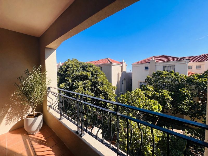 A covered balcony featuring a wrought iron balustrade and tiled flooring, offering a pleasant view of the surrounding residential complex and greenery.