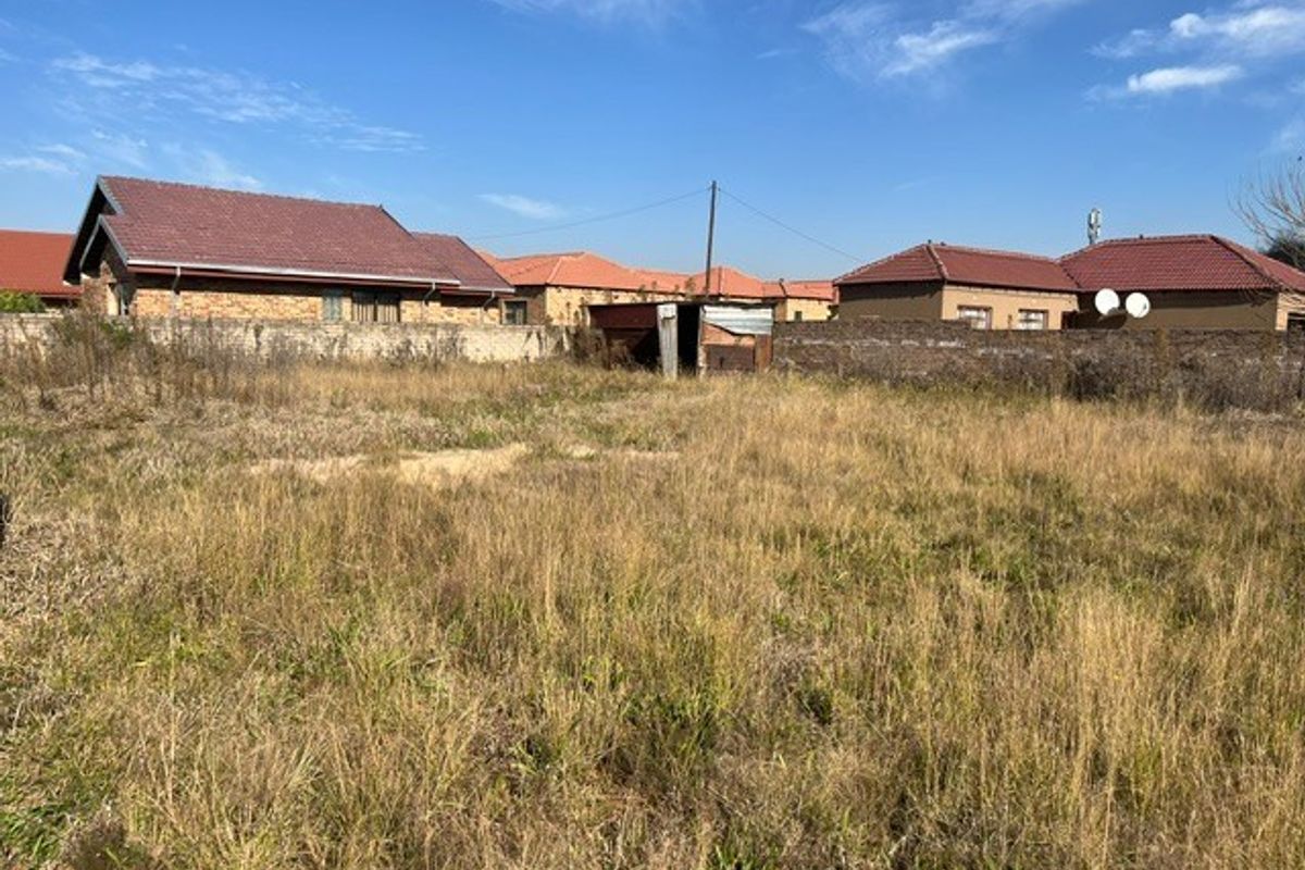 A vacant residential plot featuring overgrown grass, enclosed by perimeter boundary walls with a small dilapidated shed structure on the property.
