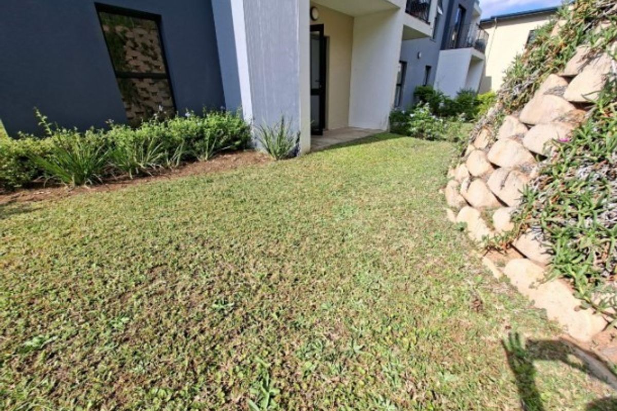 A view of the apartment's exterior rear garden area, featuring a small lawn, stone retaining wall, and access to a covered patio.