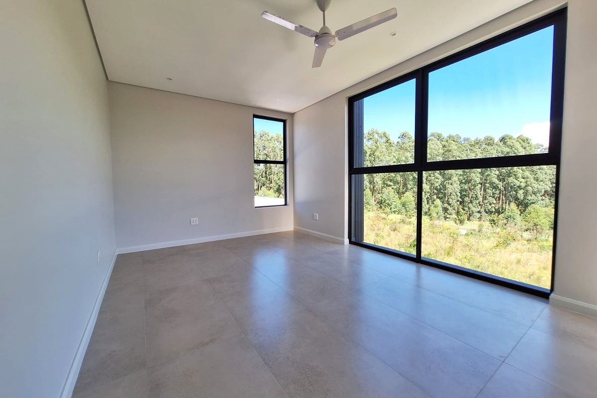 A bright, modern, empty bedroom featuring large floor-to-ceiling windows with scenic forest views, neutral tiled flooring, and a ceiling fan.
