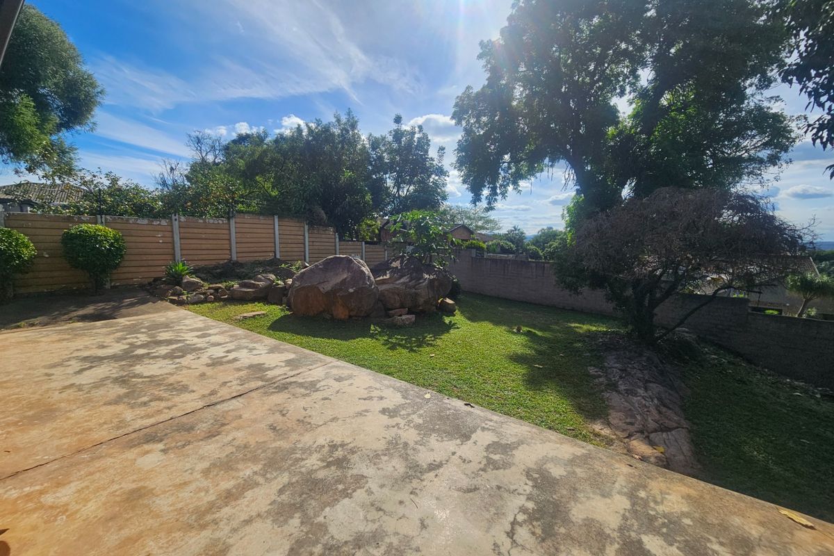 A spacious rear garden featuring a concrete patio, established trees, natural rock formations, and perimeter boundary walls.