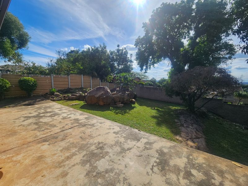 A spacious rear garden featuring a concrete patio, established trees, natural rock formations, and perimeter boundary walls.