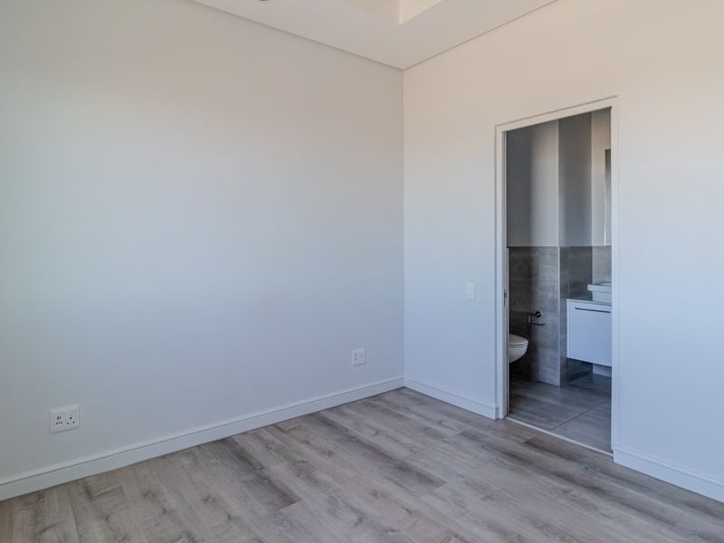 A bright, empty bedroom featuring modern wood-look flooring, neutral walls, and an adjoining en-suite bathroom with contemporary fixtures.