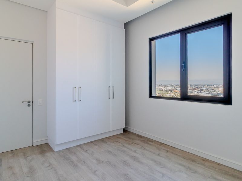 A bright, modern bedroom featuring light wood-look flooring, built-in white cabinetry, and a large window offering expansive city views.