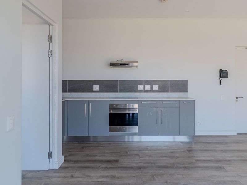 A modern, minimalist kitchen featuring grey cabinetry, a built-in oven, extractor fan, and sleek tiled backsplash, set within an open-plan living space.