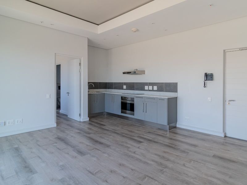 Modern open-plan apartment featuring a sleek kitchen with built-in oven, hob, and extractor fan, complemented by light wood-look flooring and recessed ceiling lighting.
