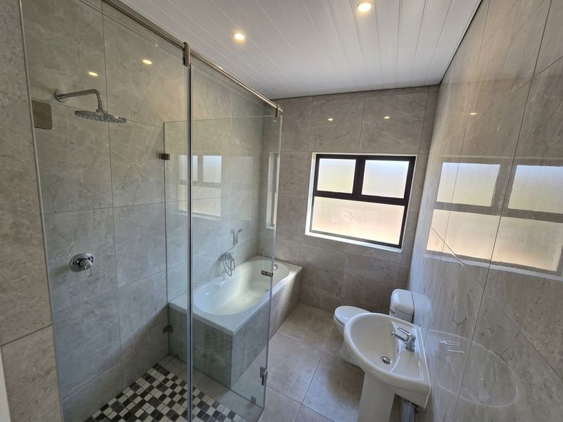 Modern bathroom featuring a glass-enclosed shower, built-in bathtub, pedestal basin, and toilet, finished with neutral floor-to-ceiling tiles and recessed lighting.