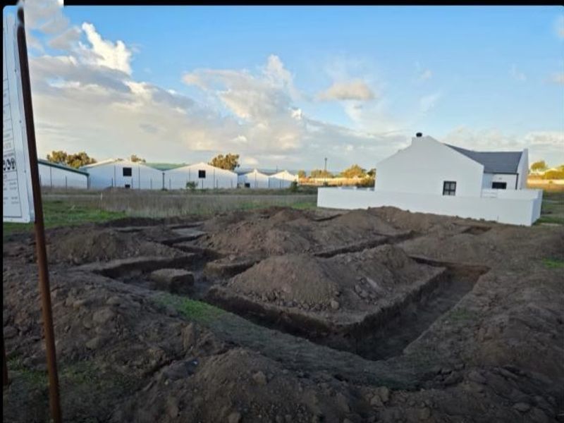 A construction site showing excavated foundation trenches for a new building project, with an existing white house visible in the background.