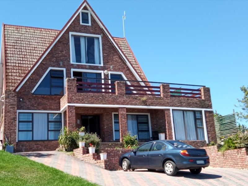 A distinctive A-frame brick house featuring a prominent upper-level balcony, large windows, and a paved driveway leading to the main entrance.